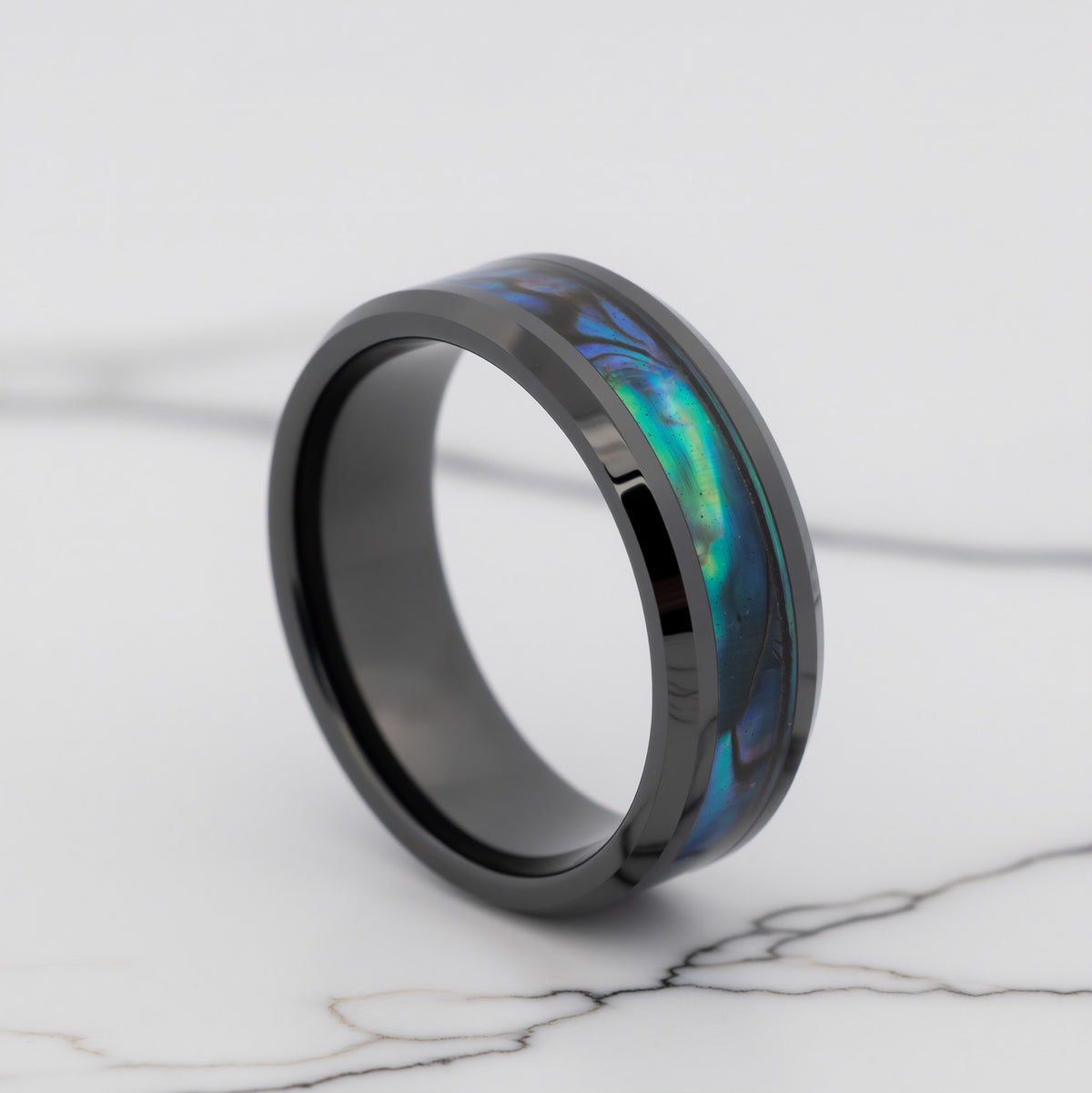 A black ring with an inlaid iridescent abalone shell segment showing blue, green, and purple hues, displayed on a white marble surface.