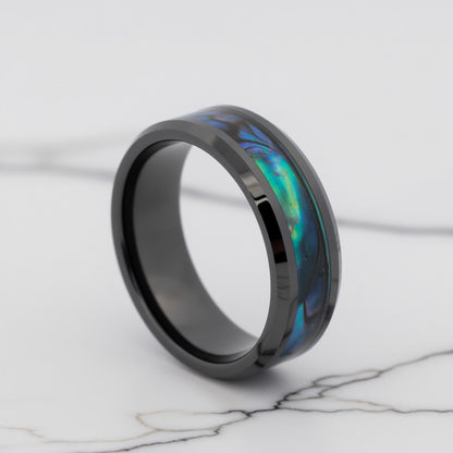 A black ring with an inlaid iridescent abalone shell segment showing blue, green, and purple hues, displayed on a white marble surface.