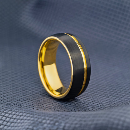 A black and gold ring with a brushed black center band and polished gold edges, resting on a textured gray fabric background.