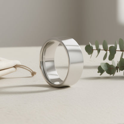 A polished silver band ring standing upright on a light beige surface, with a small white pouch on the left and green eucalyptus leaves on the right in a softly lit studio setting.
